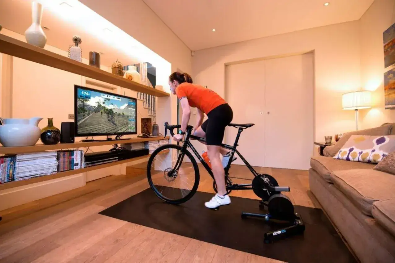 Kobieta ćwiczy na rowerze stacjonarnym przed telewizorem, wirtualnie przemierzając trasę. Sprawdź kinomap cena i zacznij trening w domu!
