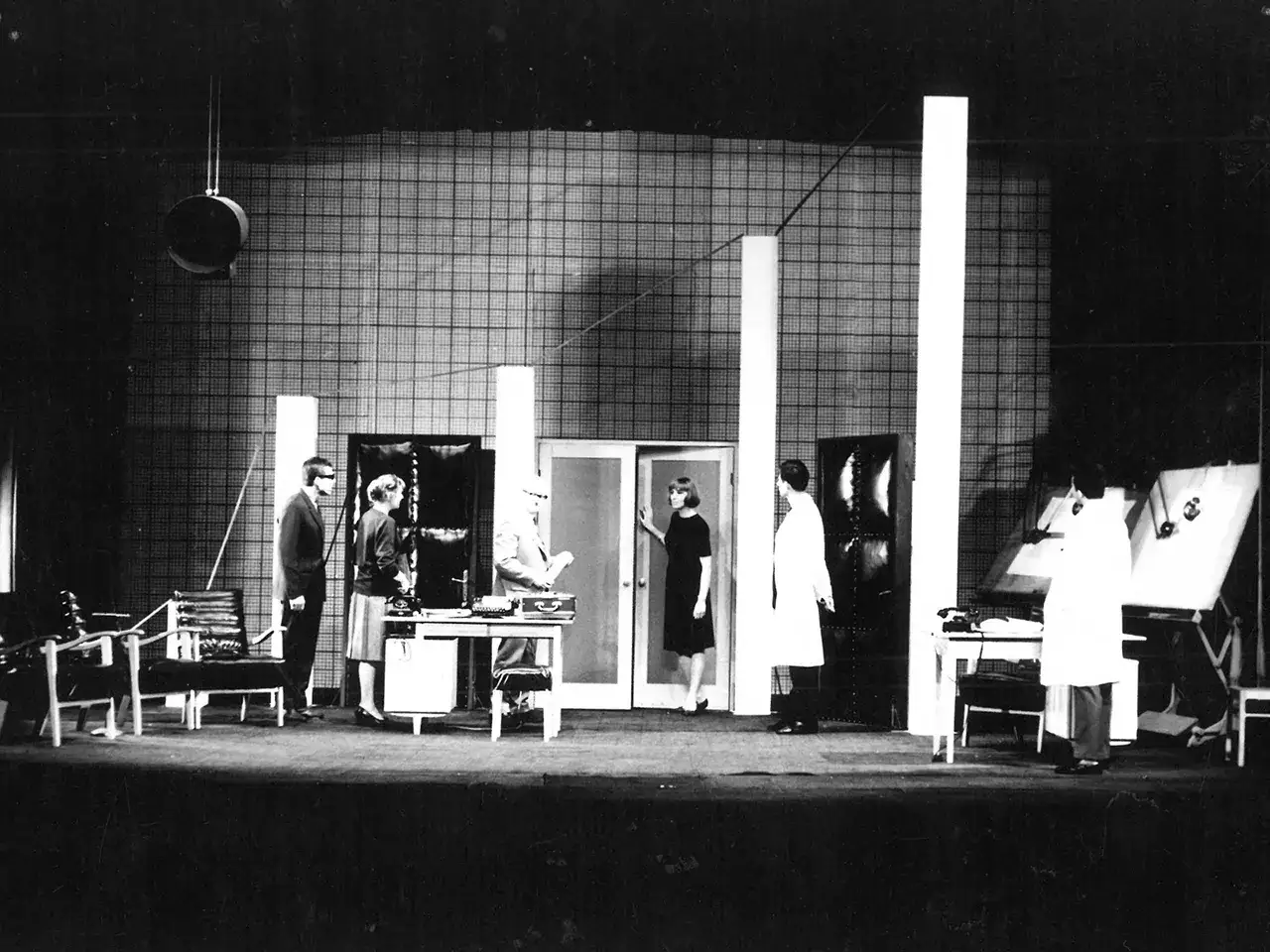 Scena teatralna z ludźmi w białych fartuchach i eleganckich strojach. To może być fragment ich tournee po świecie nauki.