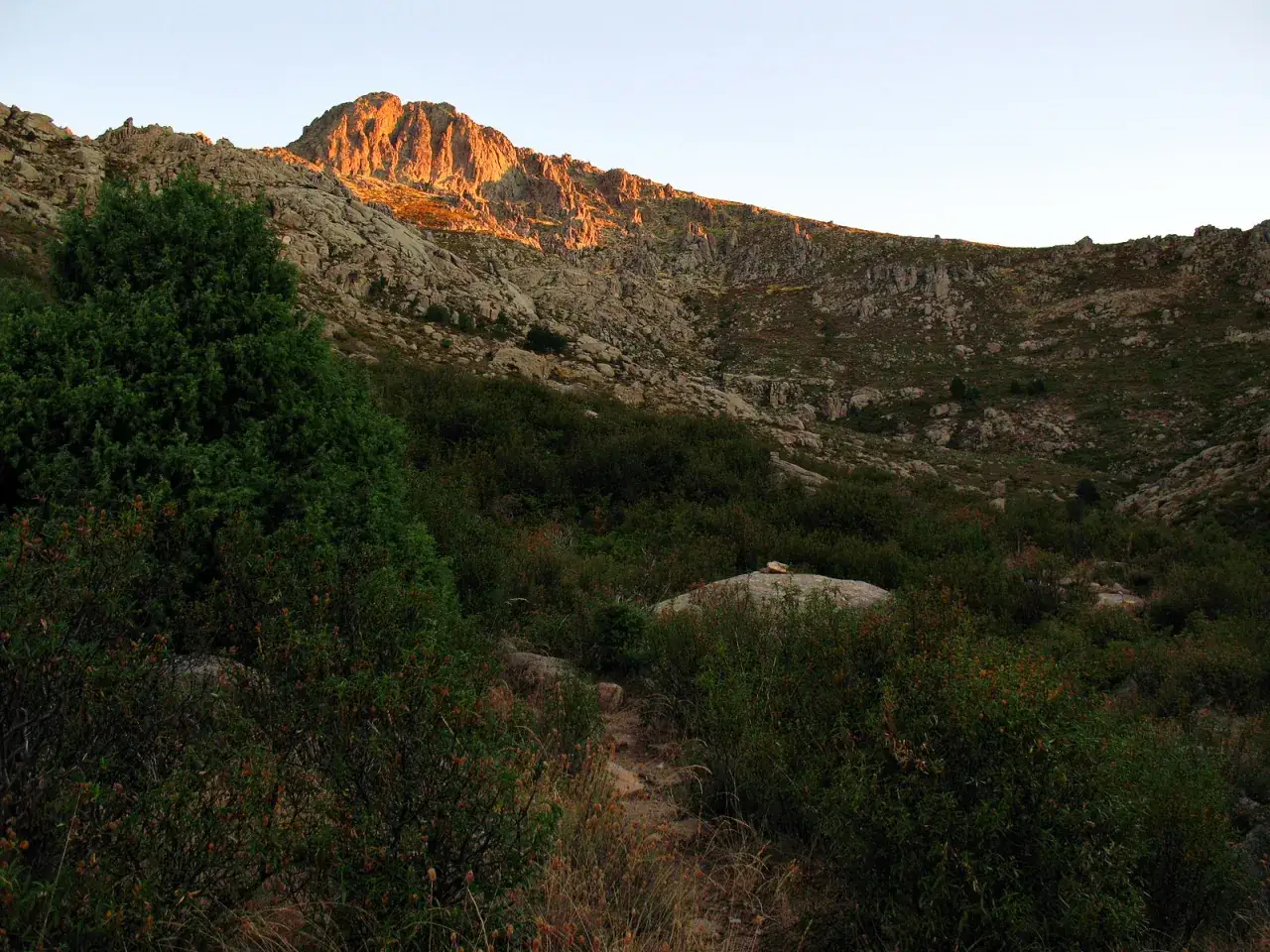 Un sendero serpentea entre matorrales y rocas hacia una montaña iluminada por el sol poniente, su cima rocosa parece la silueta de la maliciosa.