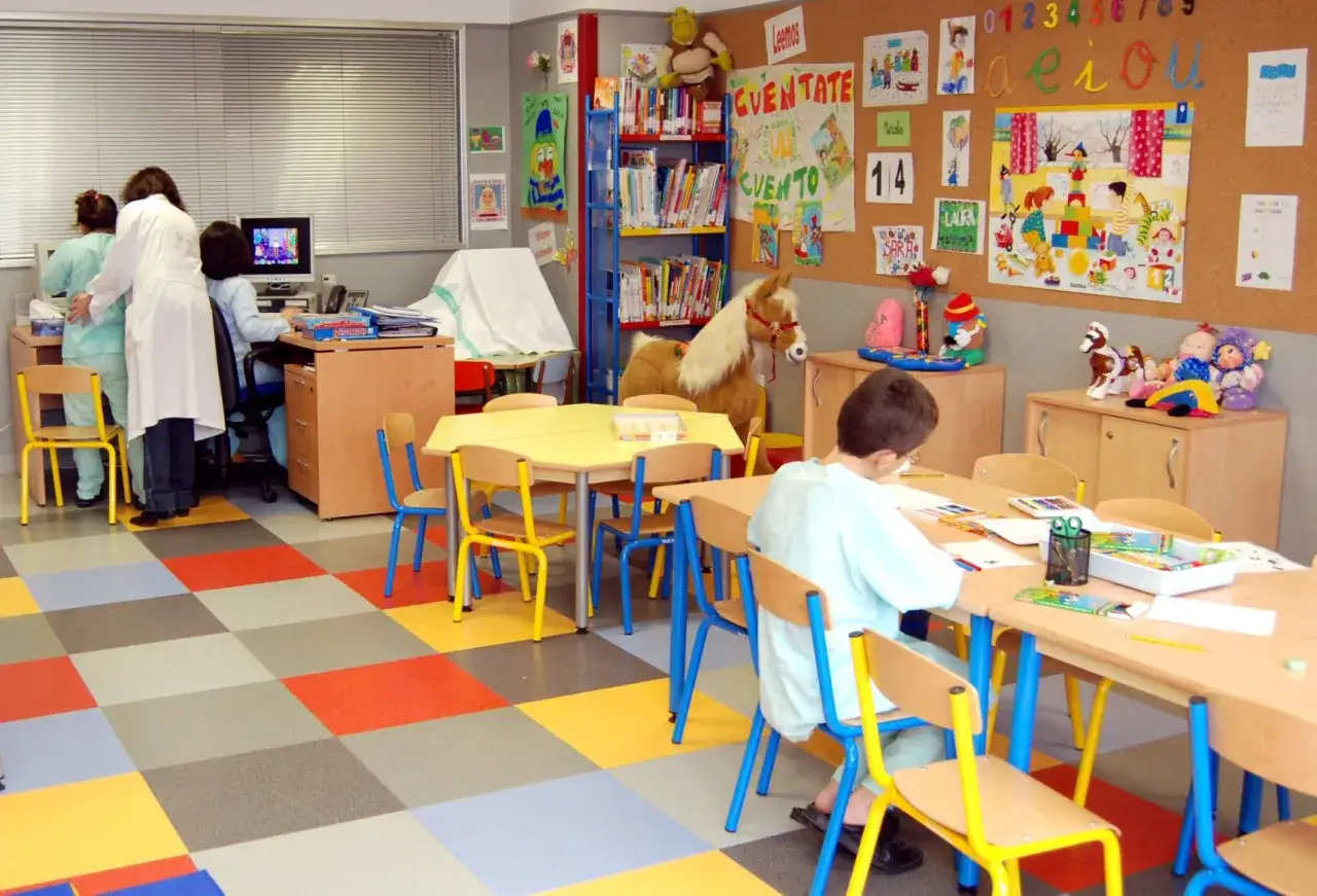 Aula infantil con estaciones de aprendizaje. Ni&ntilde;os y adultos interact&uacute;an en un espacio colorido con mesas, sillas, libros y materiales did&aacute;cticos.