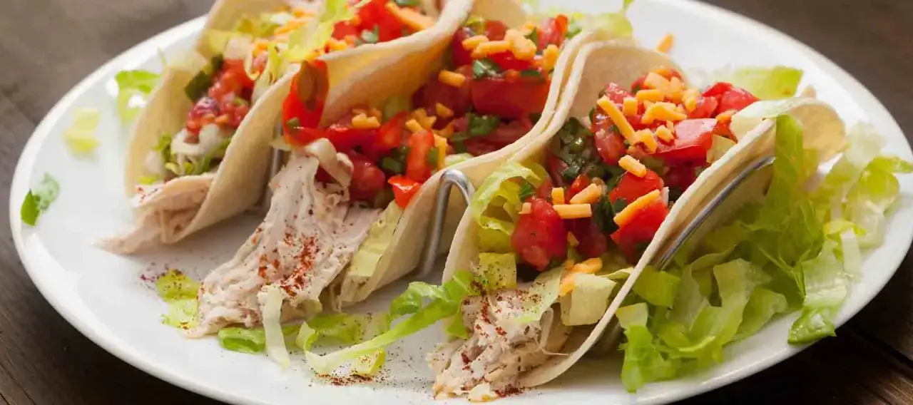 Tres tacos de pollo con lechuga, tomate picado y queso rallado sobre una tortilla suave, listos para disfrutar.