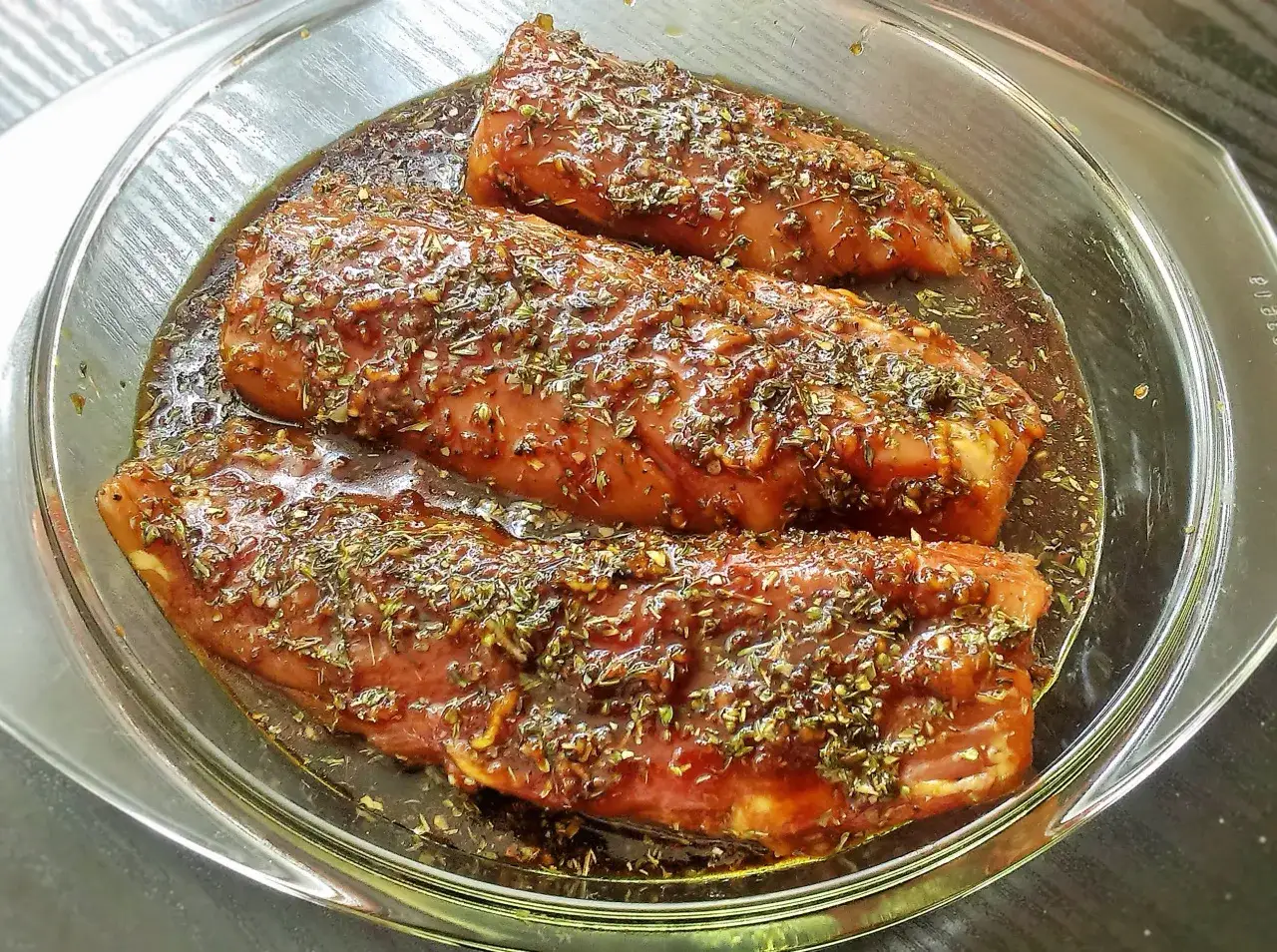 Trzy kawałki mięsa w szklanej misce, zanurzone w aromatycznej marynacie z ziołami. Idealne do grillowania, pokazują, jak zrobić marynatę do mięsa.