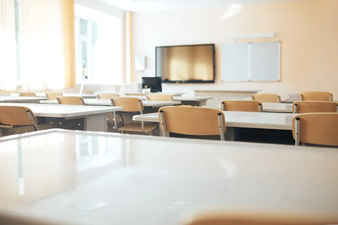 Pusta sala lekcyjna z rzędami ławek i krzeseł, gotowa na lekcje. W tle tablica interaktywna i białe tablice, które wspierają statut szkoły.