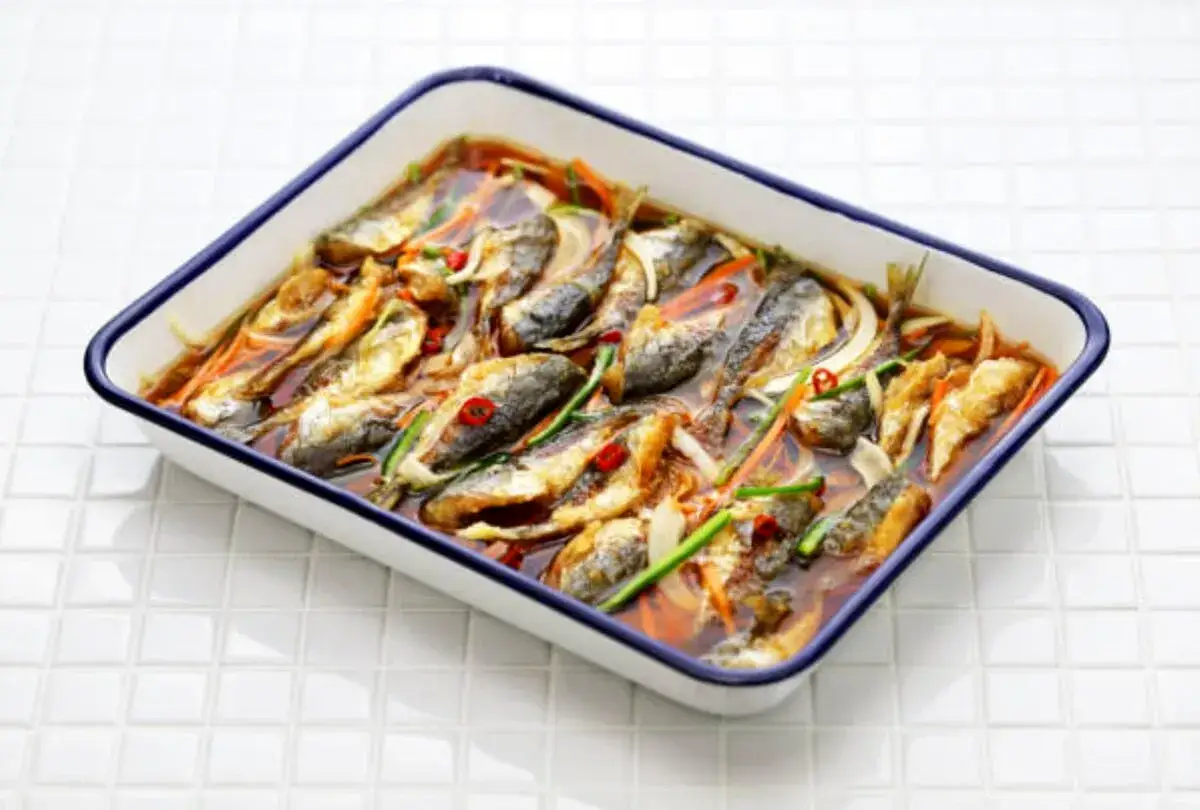 Piatto di pesce in carpione, con pesci interi disposti in una teglia bianca con bordi blu, immersi in un liquido ambrato con verdure.