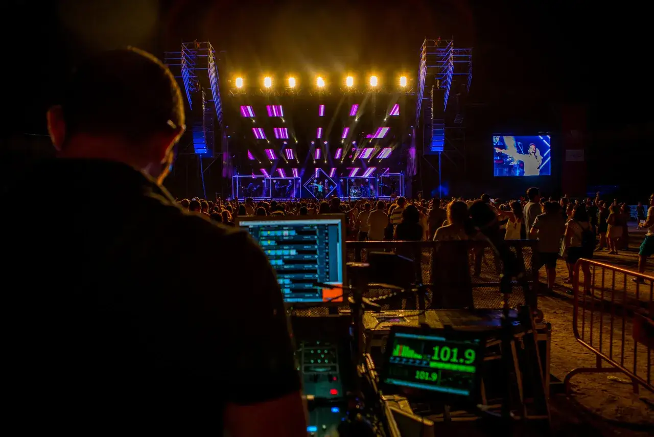 Technicien ajustant le son d'un concert, &eacute;cran affichant la mesure decibel. Foule en d&eacute;lire devant la sc&egrave;ne illumin&eacute;e.
