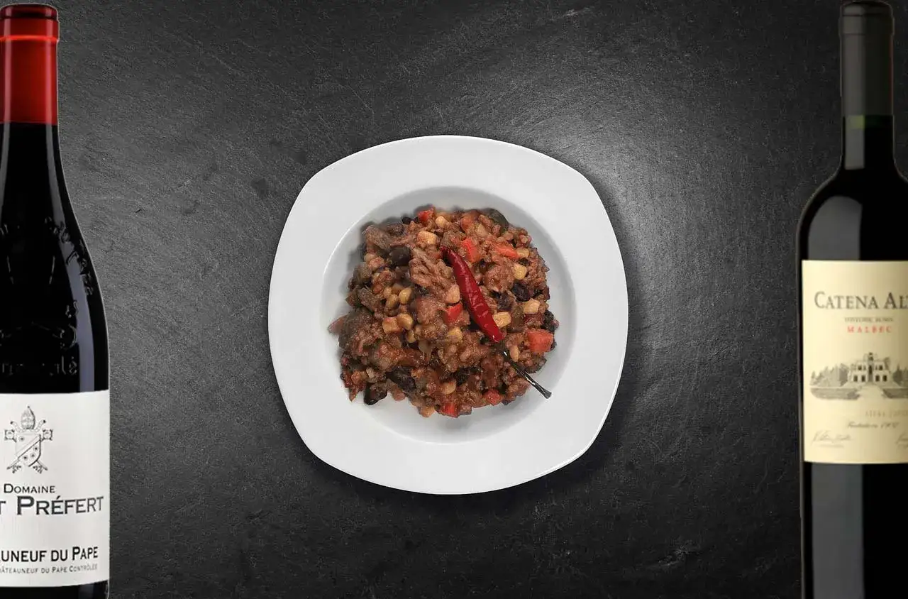 En tallrik med chili con carne, toppad med en röd chili, flankerad av två flaskor rött vin. Perfekt vin till chili con carne.