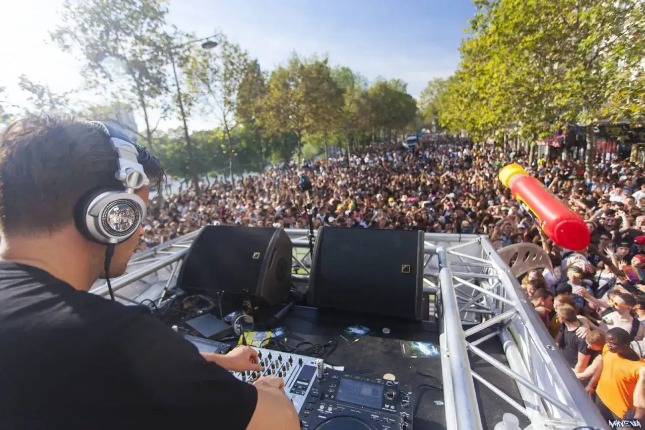Un DJ à la mode mixe devant une foule immense, sous un ciel bleu. Une ambiance de fête en plein air.