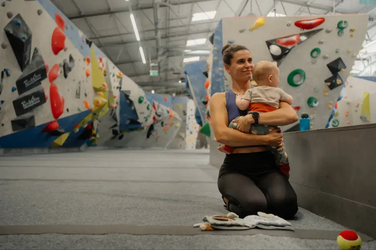 Eine Frau mit Baby in den Armen in einer Kletterhalle. Sie scheint sich auf das Klettern vorzubereiten, vielleicht nach der Schwangerschaft.