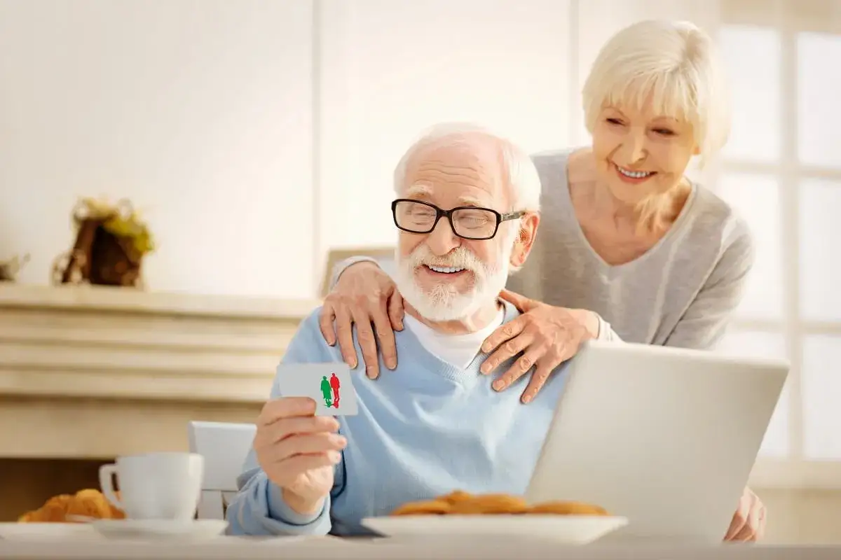 Szczęśliwa para seniorów przegląda ofertę bezpłatnych badań po 50. na laptopie, trzymając kartę z symbolami ludzi.