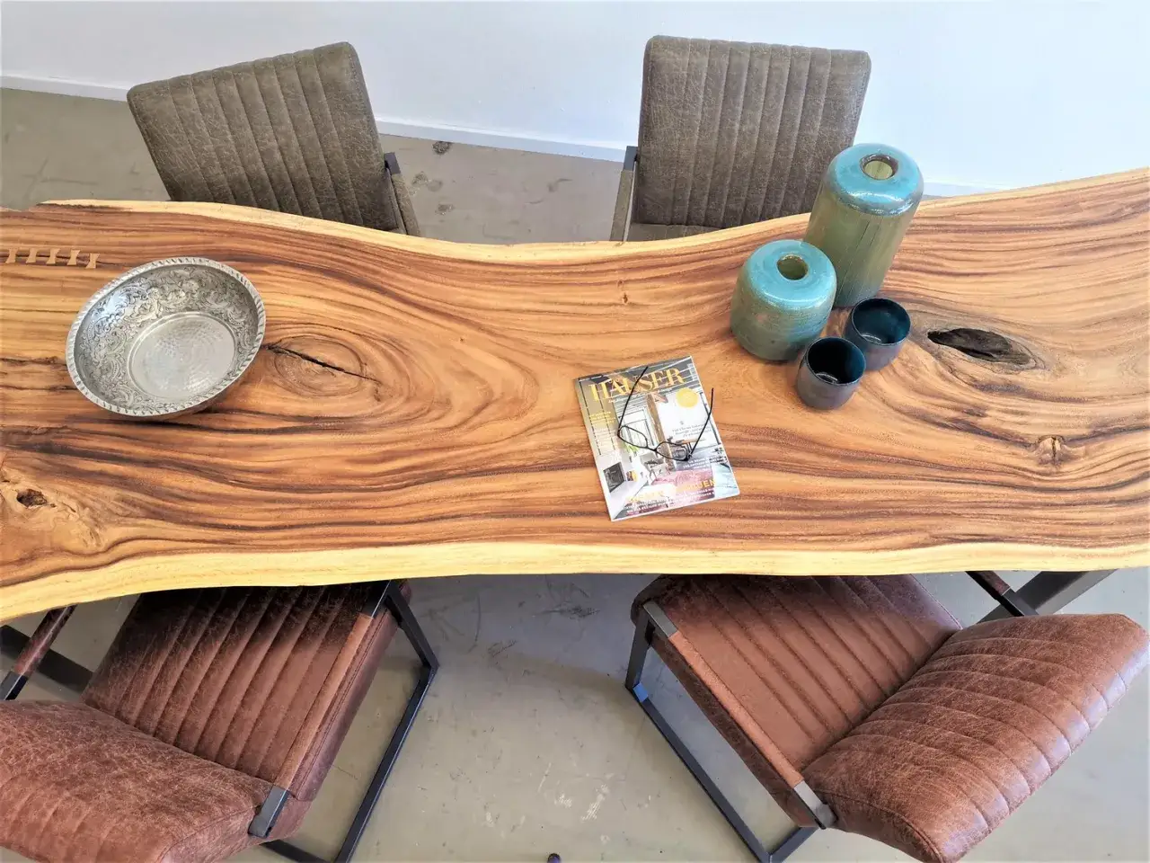 Ein massiver Holztisch mit natürlicher Kante, dekoriert mit Vasen und einer Schale. Lederstühle umgeben den Tisch.