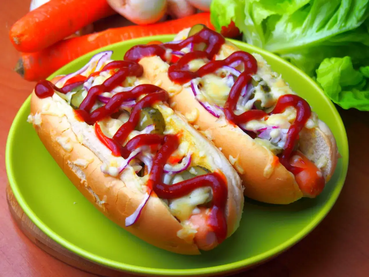 Dwa pyszne, domowe hot dogi z serem, ogórkiem i czerwoną cebulą, polane ketchupem.