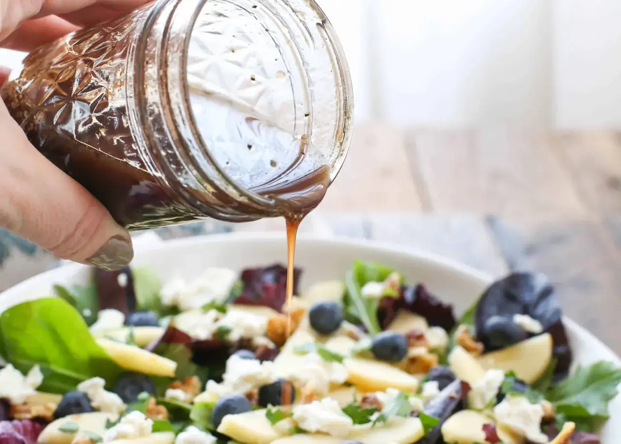 Ein Glas mit Balsamico-Dressing wird &uuml;ber einen bunten Salat mit Blaubeeren, &Auml;pfeln und K&auml;se gegossen.