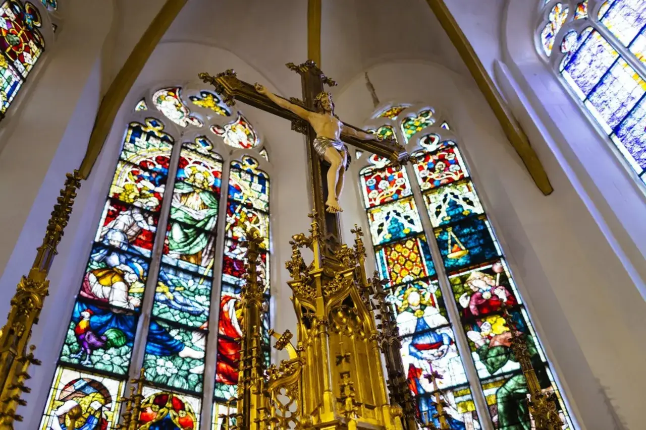 Ein goldenes Kreuz mit Jesusfigur vor farbenprächtigen Glasfenstern. 