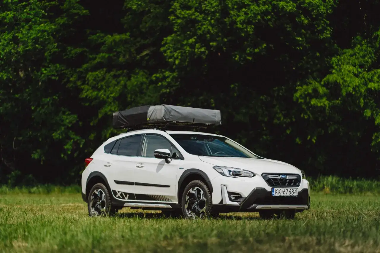 Biały Subaru XV z namiotem dachowym na tle zielonego lasu. Wiele pozytywnych subaru xv opinie potwierdza jego wszechstronność.