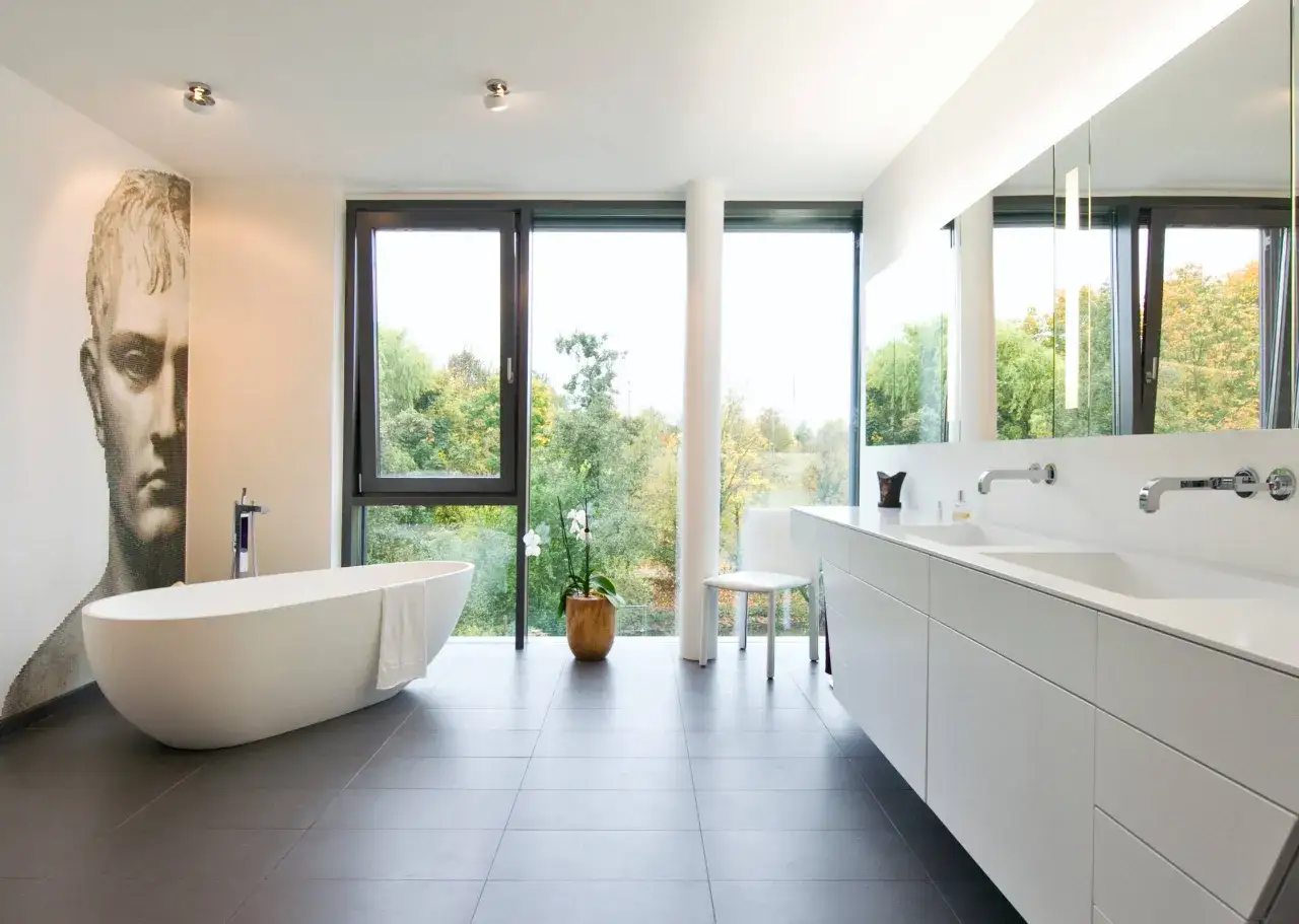 Eine freistehende **Badewanne mitten im Raum** mit Blick ins Grüne. Daneben ein Doppelwaschbecken und ein Porträt an der Wand.