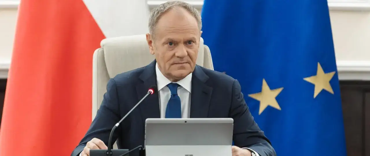 Donald Tusk, premier Polski, siedzi przy stole z flagą UE i Polski. Jego mina sugeruje, że może być gotów nałożyć **veto**.