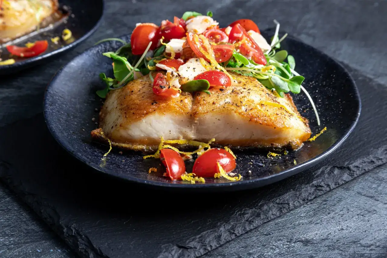 Smażony filet z dorsza czarnego, podany z pomidorkami koktajlowymi, kiełkami i skórką cytrynową na czarnym talerzu.