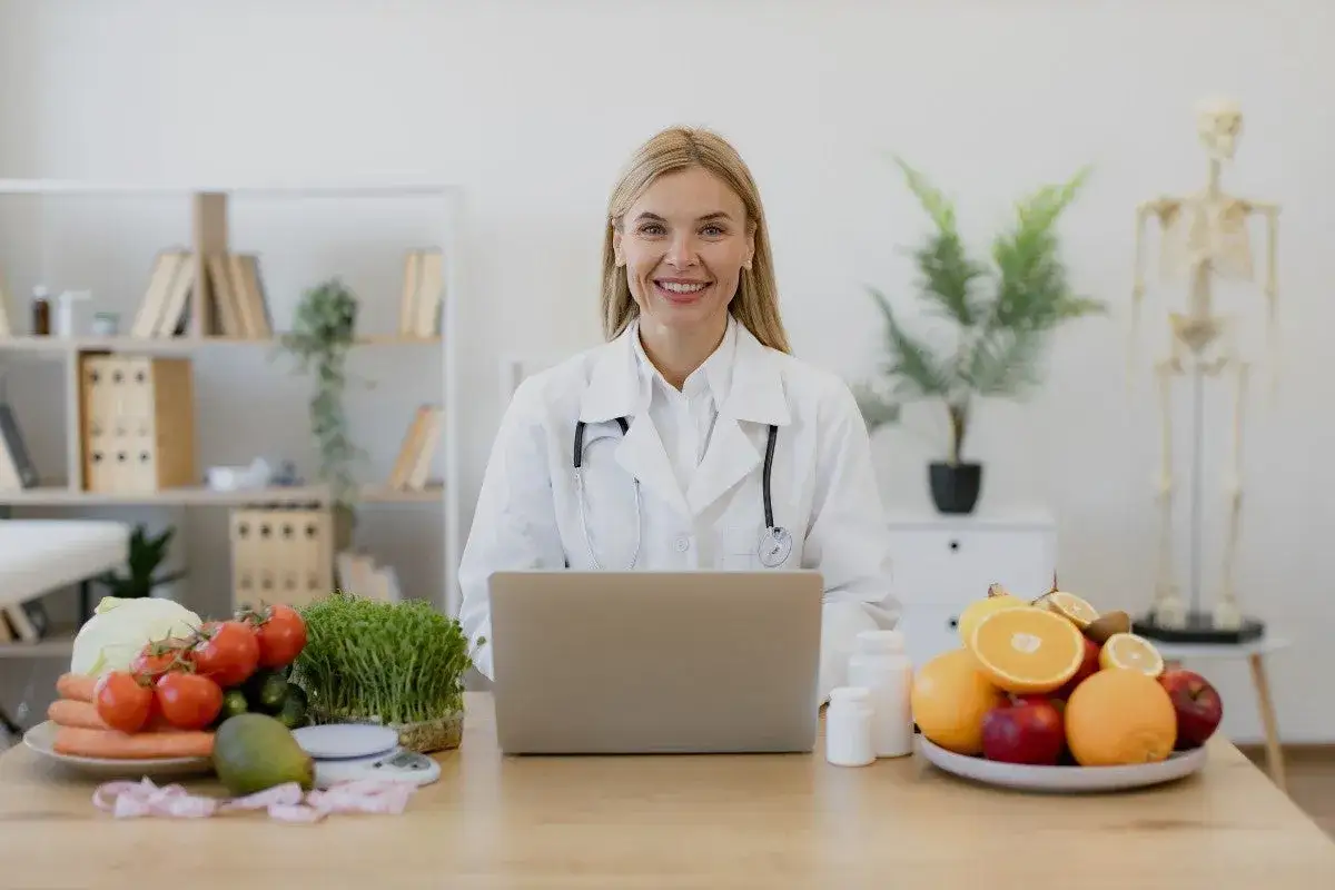 Nutricionista sonriente con frutas y verduras, lista para tu consulta de diet&eacute;tica a distancia.