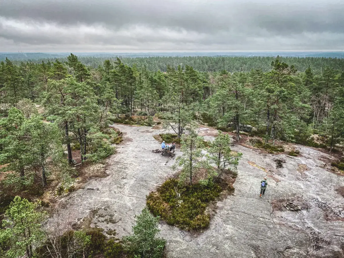 Vacker utsikt &ouml;ver paradiset naturreservat med skog och klippor. Tv&aring; personer sitter vid ett bord, en vandrare g&aring;r p&aring; klippan.