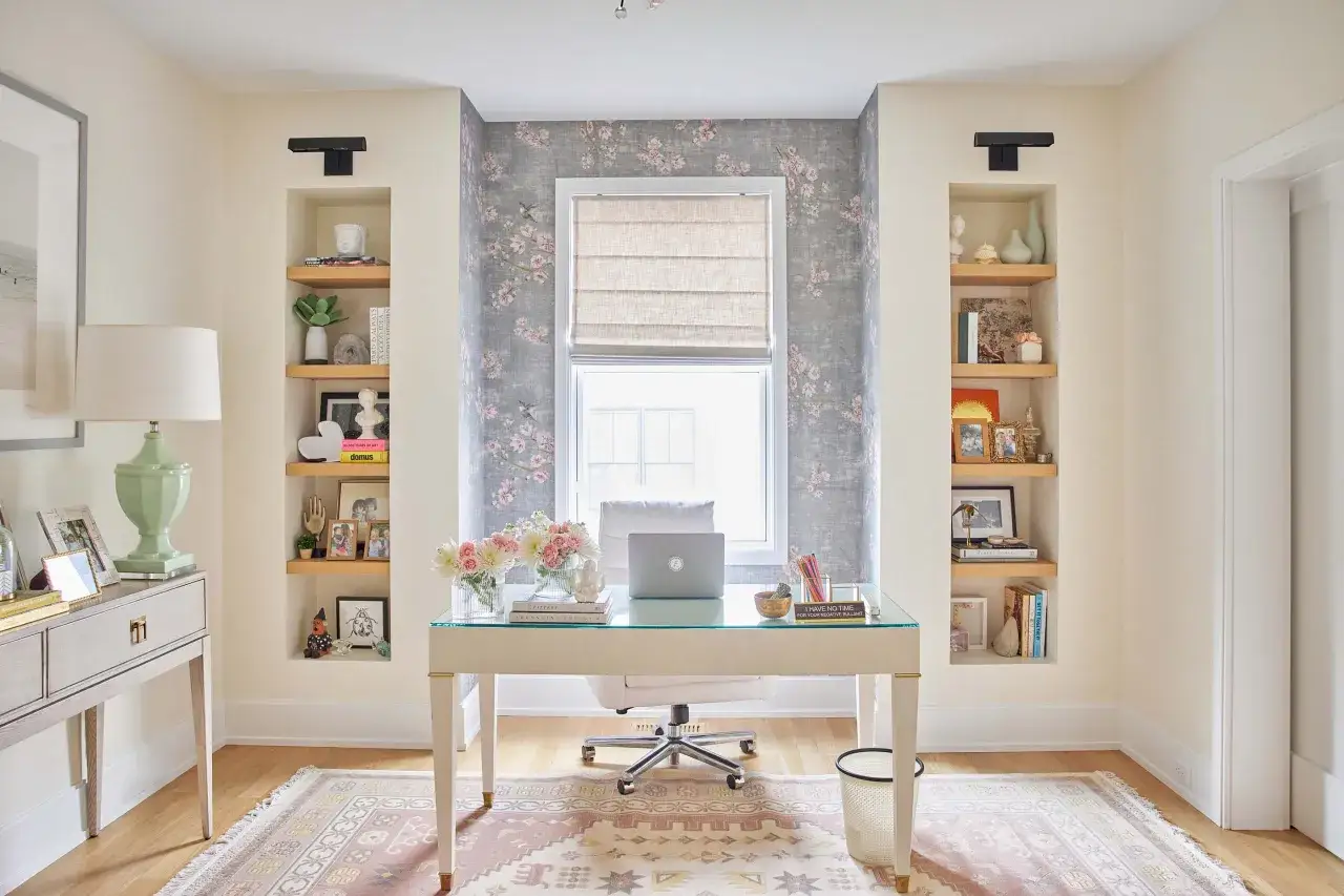 Bureau lumineux avec papier peint floral, bureau en verre, ordinateur portable et étagères décorées. La **couleur** douce et les objets choisis favorisent la **concentration**.
