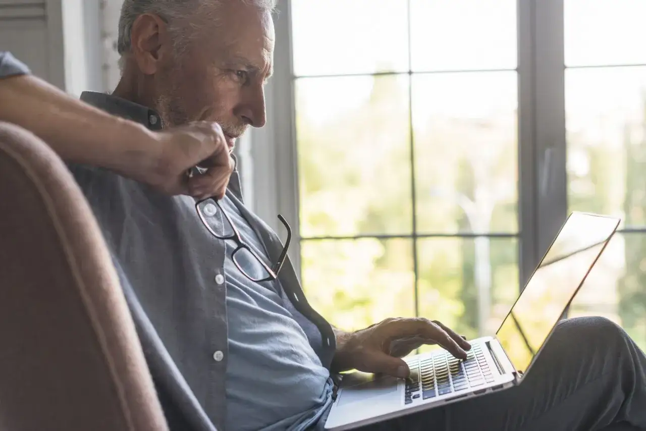 Starszy mężczyzna z okularami na koszuli pracuje na laptopie, planując swoją pomostówkę – świadczenie przedemerytalne.