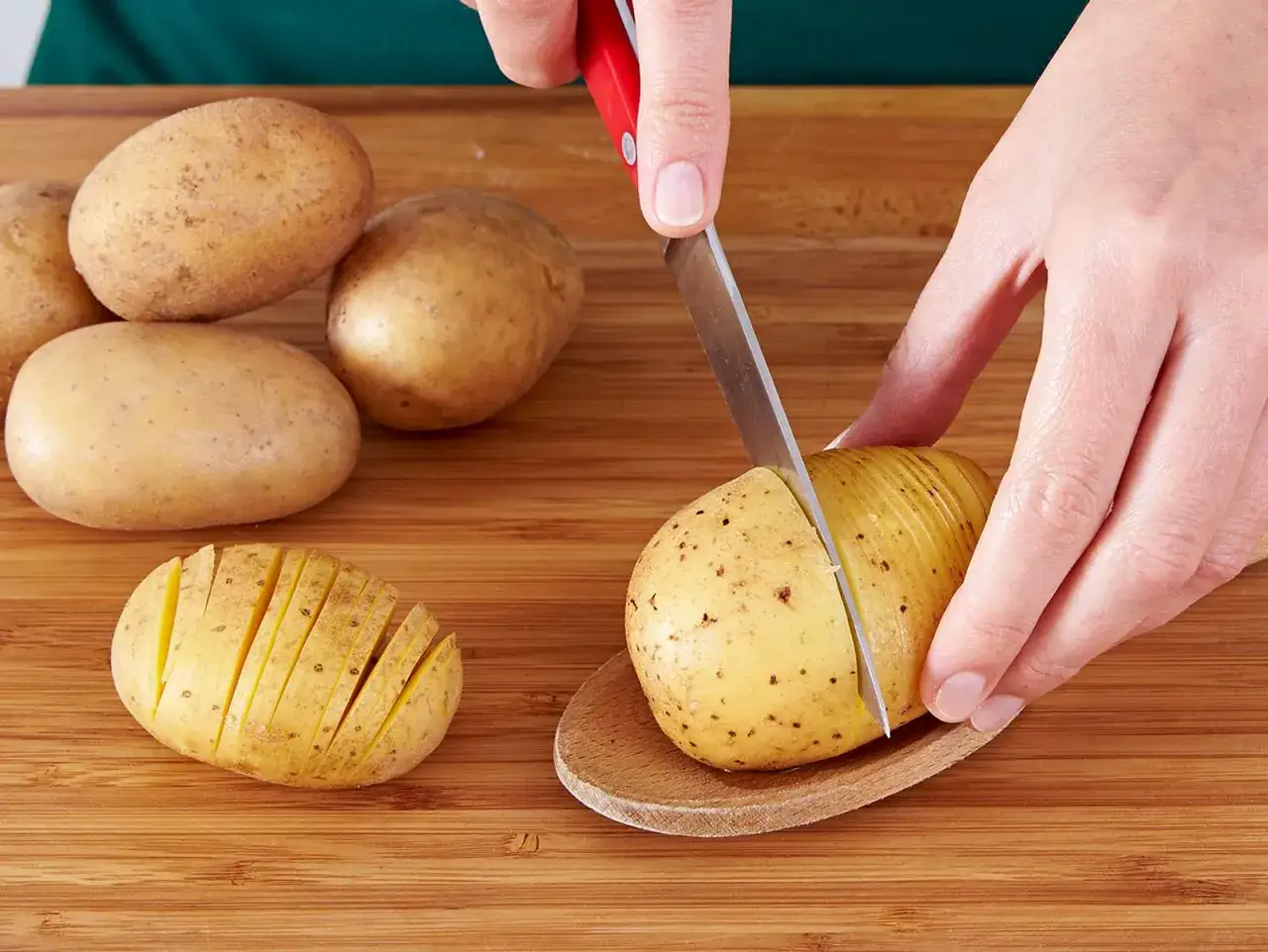 Hasselback Kartoffeln werden vorbereitet: Eine Kartoffel wird auf einem Holzlöffel liegend mit einem Messer eingeschnitten.