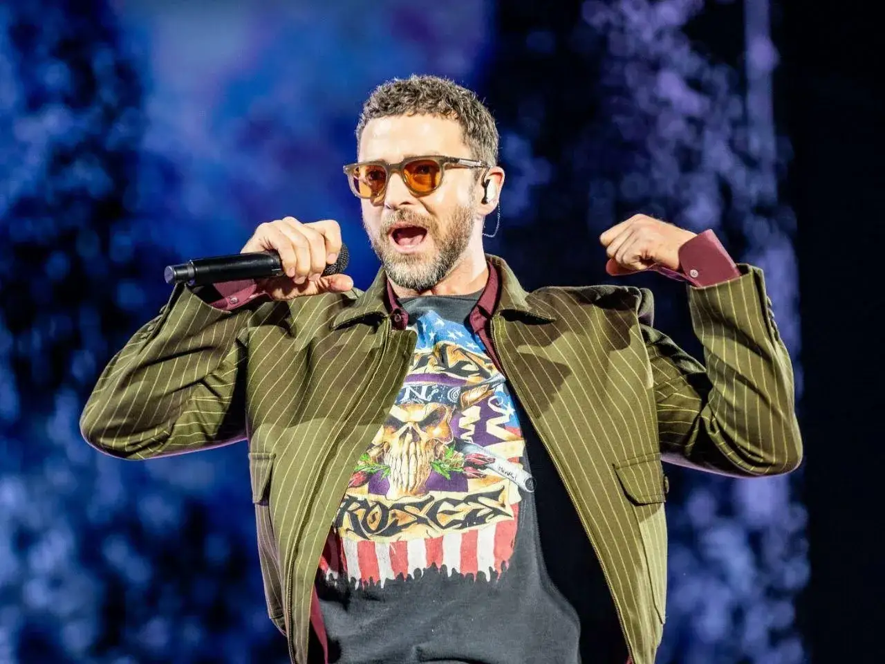 Justin Timberlake na koncercie w Polsce, w okularach i z mikrofonem w dłoni, w paski marynarce i koszulce z nadrukiem.
