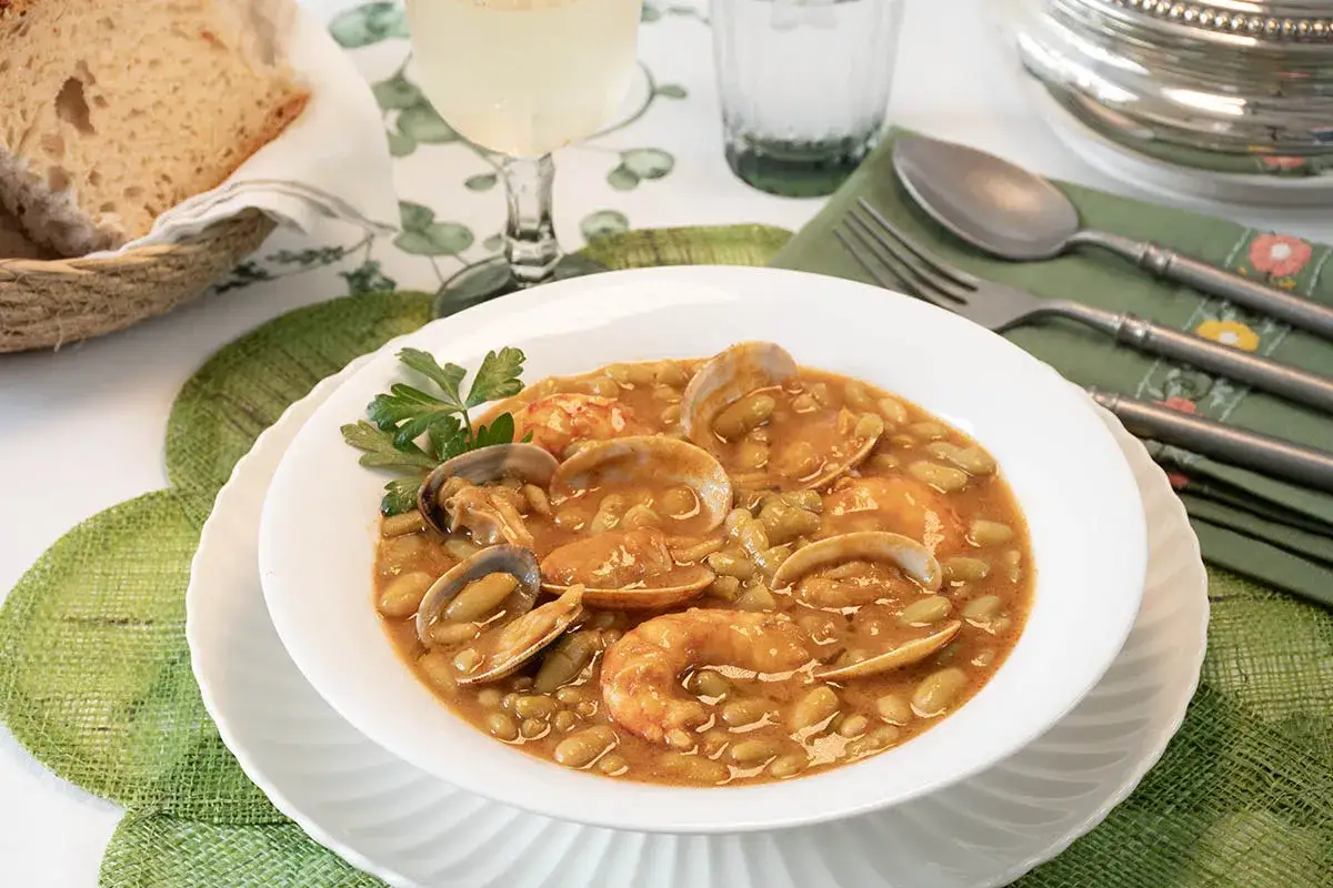 Plato de verdinas con langostinos y almejas, servido en un cuenco blanco, acompañado de pan y vino.