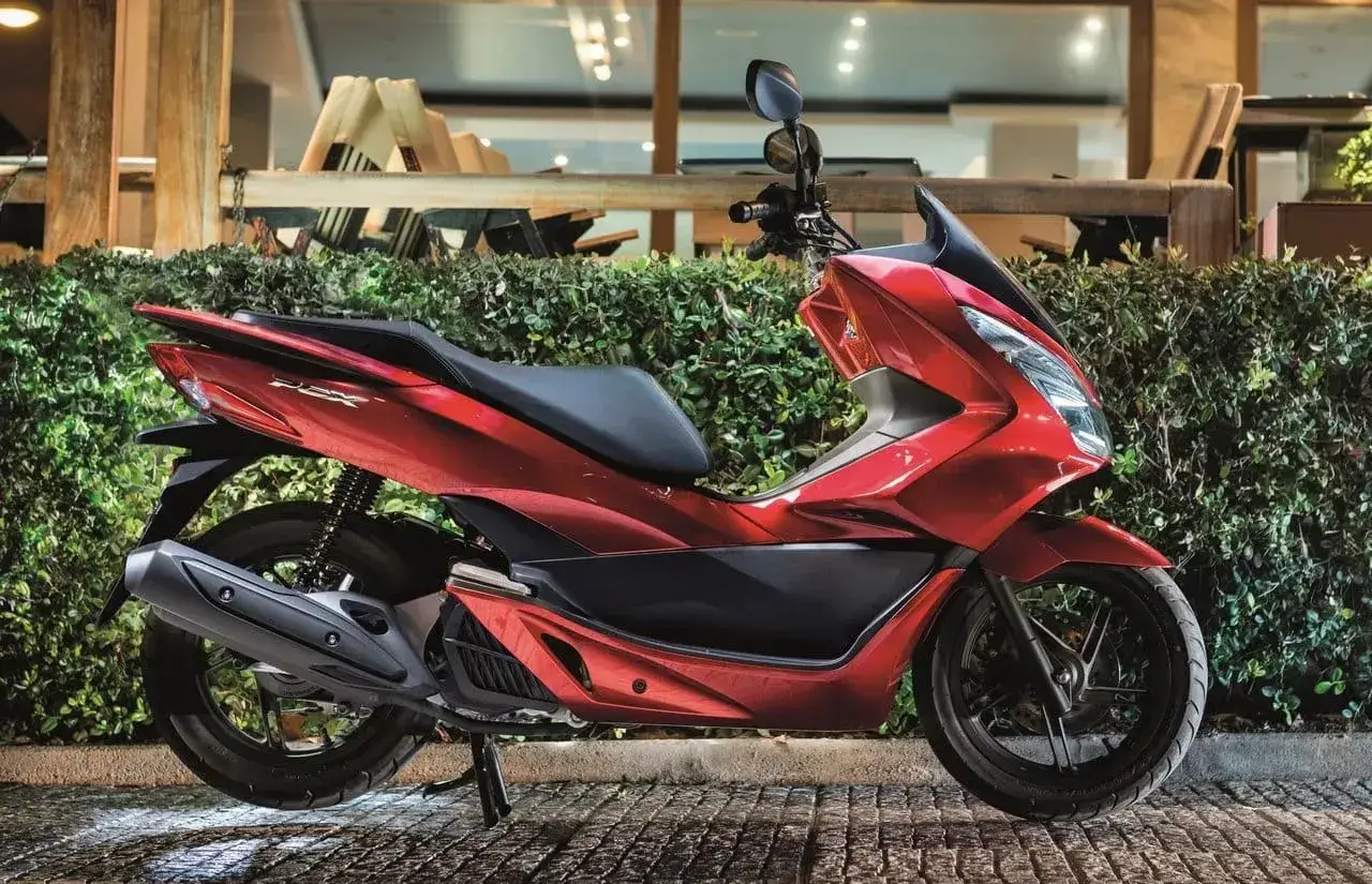Lśniący czerwony skuter Honda PCX, jeden z popularnych **marek skuterów**, zaparkowany przed zielonym żywopłotem.