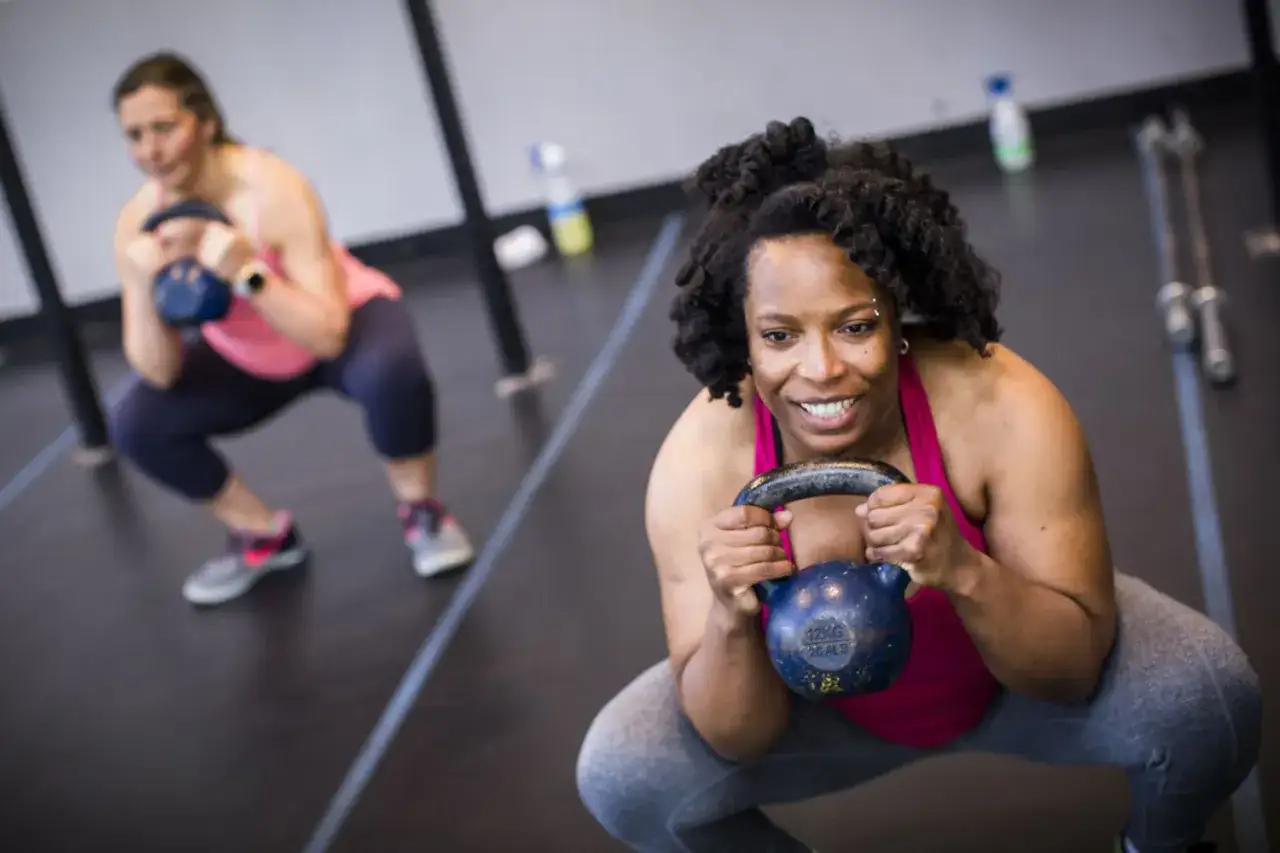 Två kvinnor gör knäböj med kettlebells. Kvinnan i förgrunden ler glatt medan hon håller en blå kettlebell.