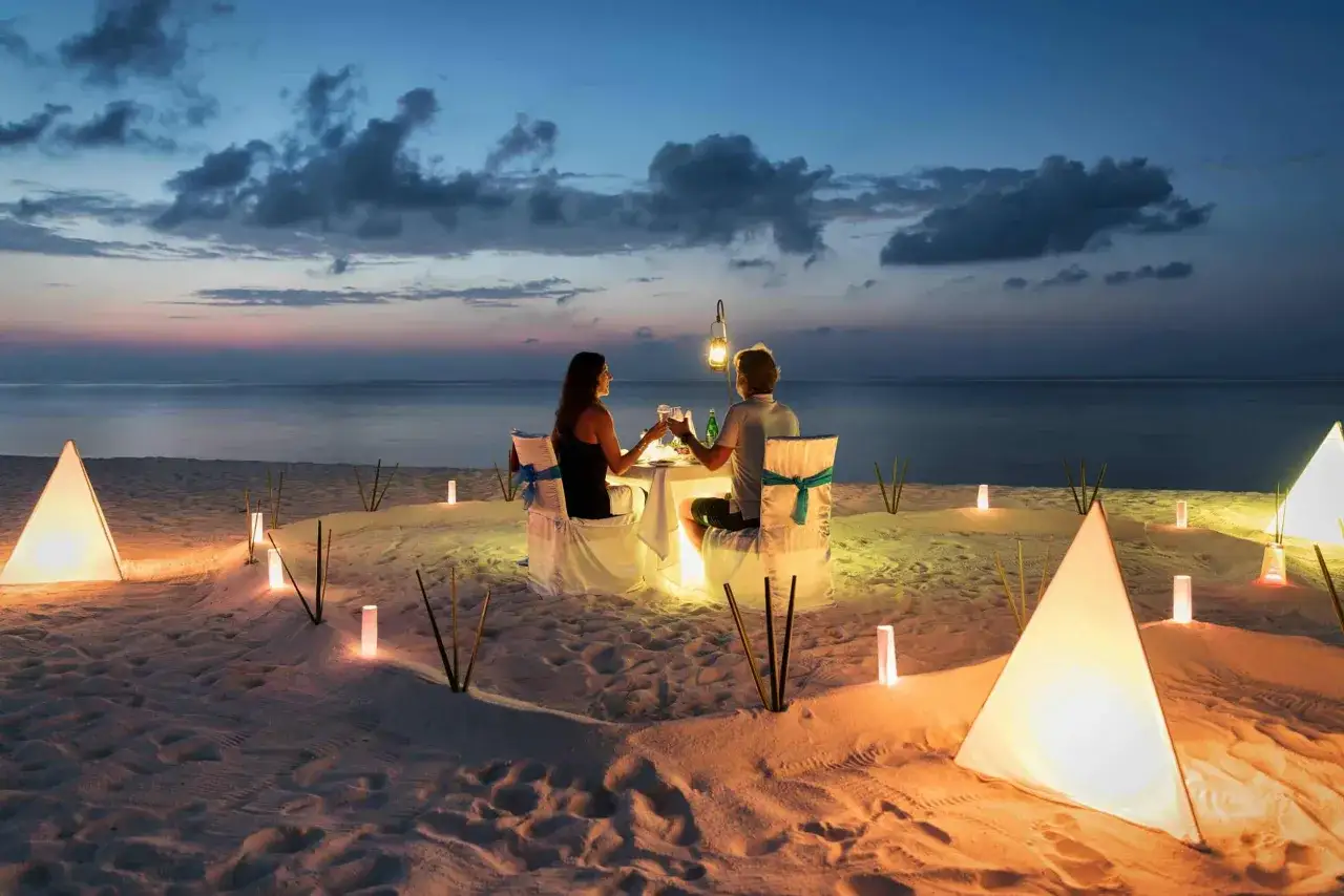 Un couple trinque lors d'un d&icirc;ner romantique sur la plage, &eacute;clair&eacute; par des lanternes. Une parfaite destination lune de miel pas cher.