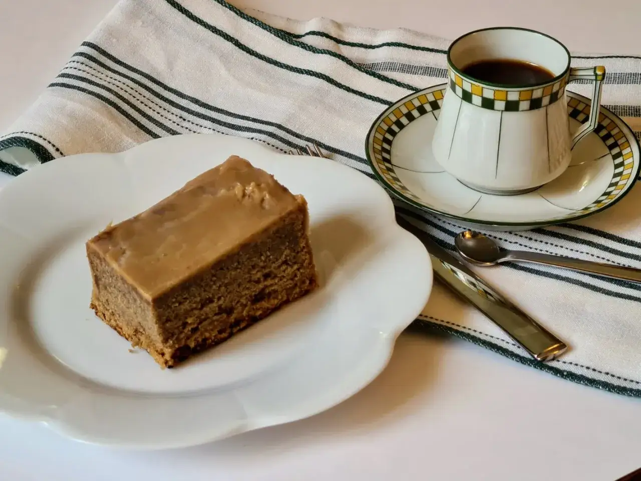 Tranche de g&acirc;teau au caf&eacute; glac&eacute; sur une assiette blanche, avec une tasse de caf&eacute; noir et des couverts sur une serviette ray&eacute;e.