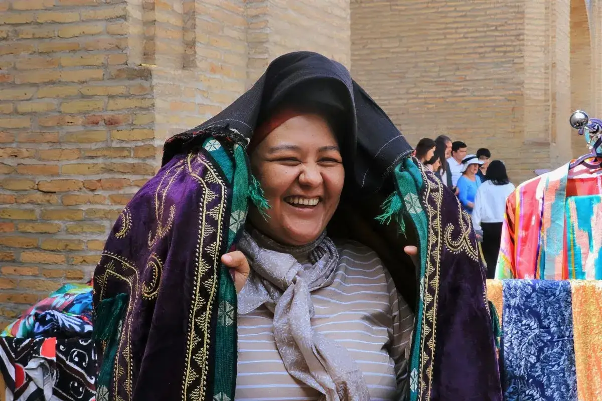 Uśmiechnięta kobieta w tradycyjnym stroju, otoczona kolorowymi tkaninami. Czy Uzbekistan jest bezpieczny? Tak, podróżni czują się tu mile widziani.
