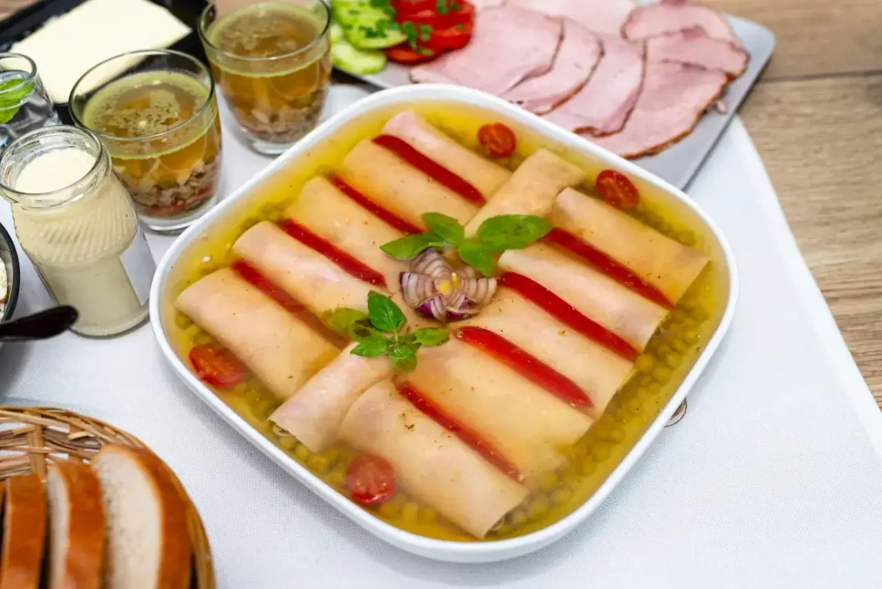 Roladki z szynki w galarecie, ozdobione papryką i ziołami, podane z pieczywem i wędliną.