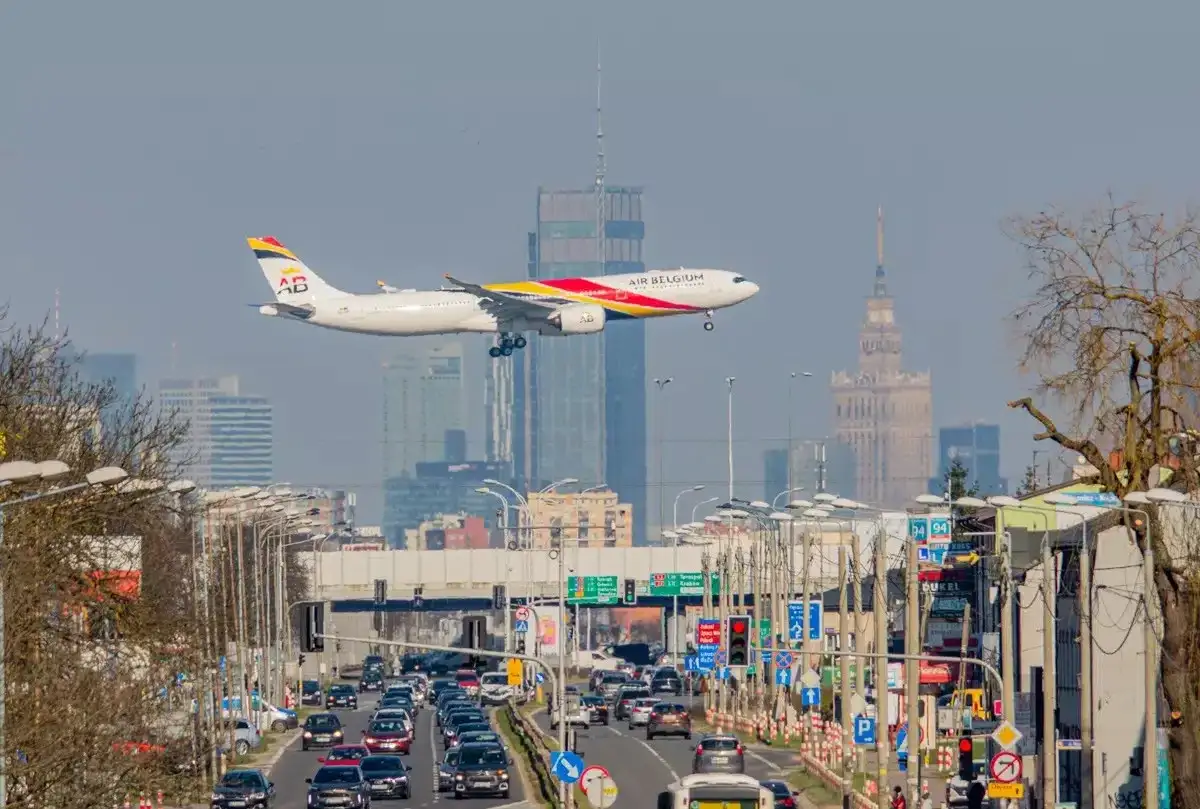Samolot Air Belgium przelatuje nad zatłoczoną trasą w Warszawie, zbliżając się do lądowania. Widok na wieżowce i Pałac Kultury.