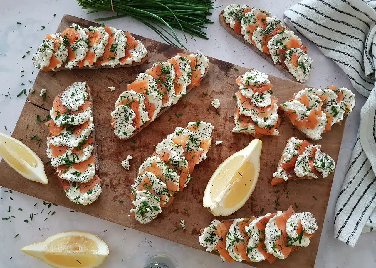 Plusieurs bruschettas saumon garnies de fromage frais, ciboulette et graines de pavot, dispos&eacute;es sur une planche en bois avec des quartiers de citron.