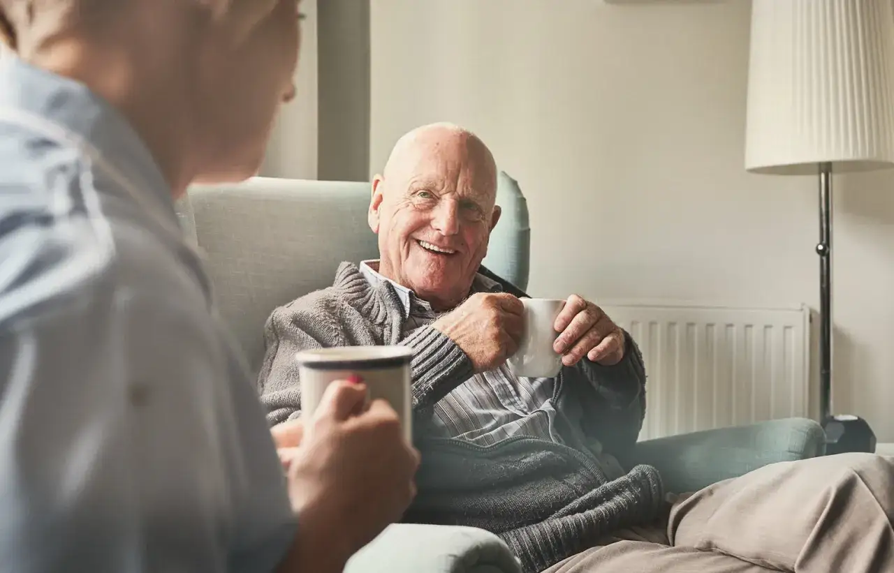Ein &auml;lterer Mann l&auml;chelt und h&auml;lt eine Tasse Kaffee. Er genie&szlig;t die Gesellschaft und die **niedrigschwelligen angebote** der Pflege.
