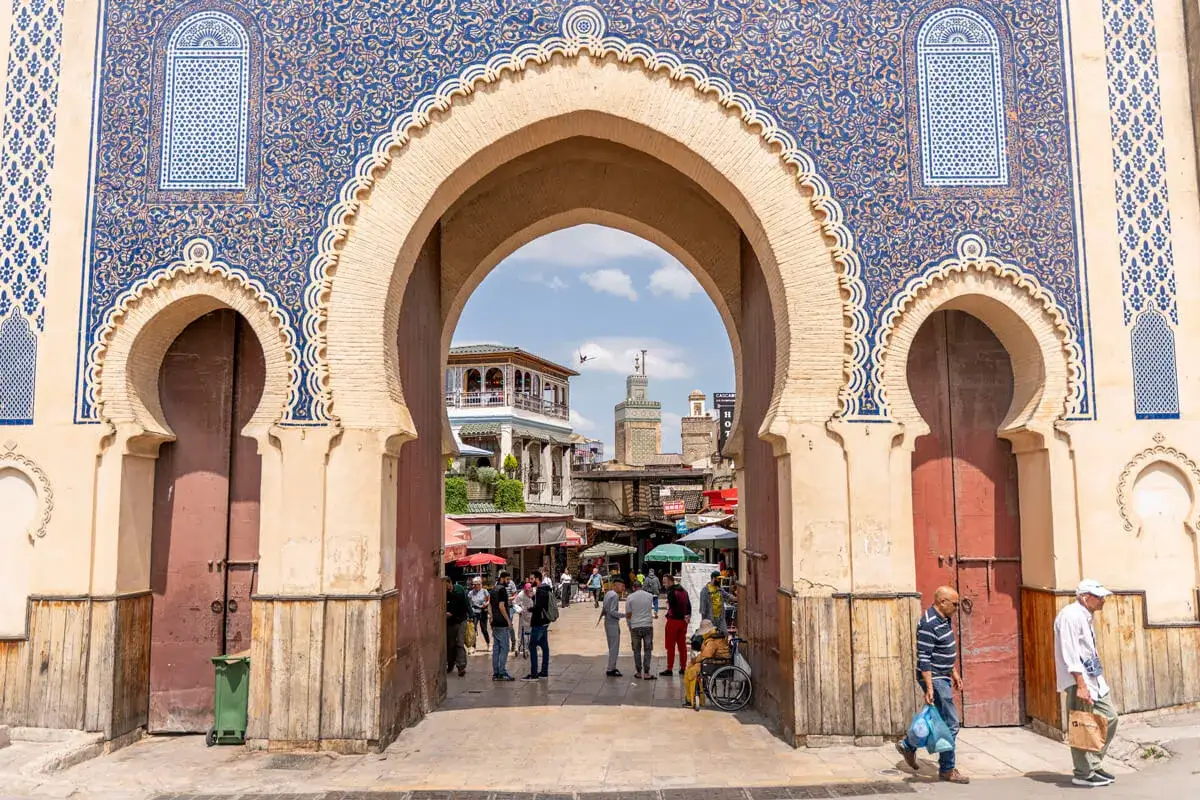 Prachtvolles Tor in Fes Stadt mit blau-goldenen Verzierungen. Menschen beleben den Platz dahinter.