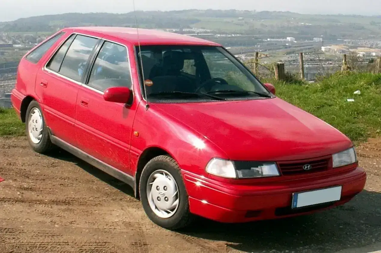 Czerwony Hyundai Pony z lat 90. stoi na błotnistej drodze, z widokiem na dolinę i wzgórza w tle.