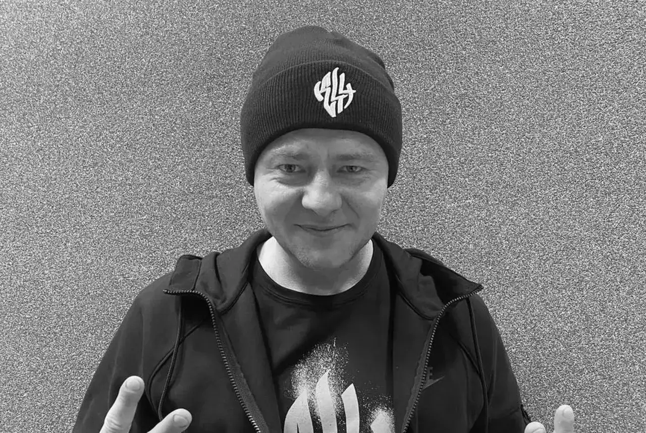 Mężczyzna w czapce z logo, w bluzie i koszulce. Nie wiadomo, **joka na co zmarł**, ale jego uśmiech jest widoczny.