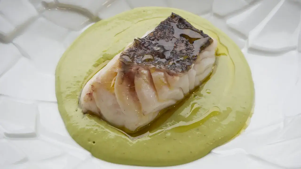 Un trozo de merluza al pil pil, con piel crujiente, sobre una crema verde en un plato blanco.