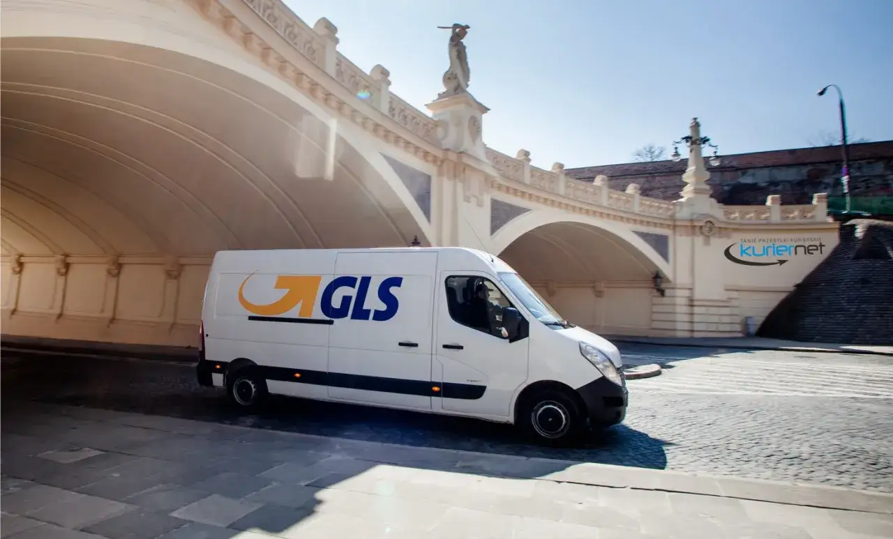Biały van GLS z logo firmy jedzie pod łukiem mostu. W tle widoczny napis 