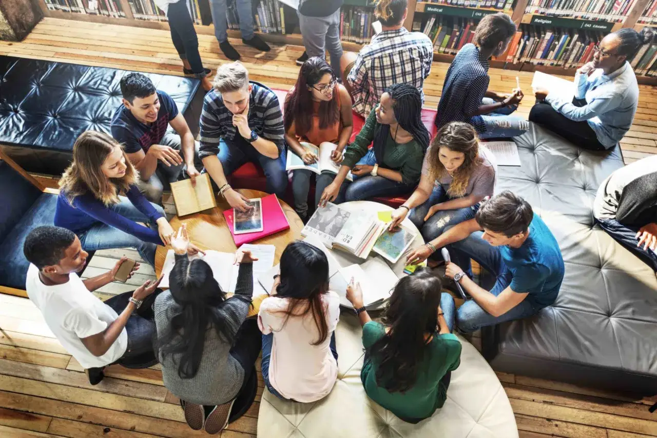 Grupa studentów dyskutuje nad materiałami, stosując metodę jigsaw. Wszyscy skupieni na książkach i notatkach, tworząc atmosferę wspólnej nauki.