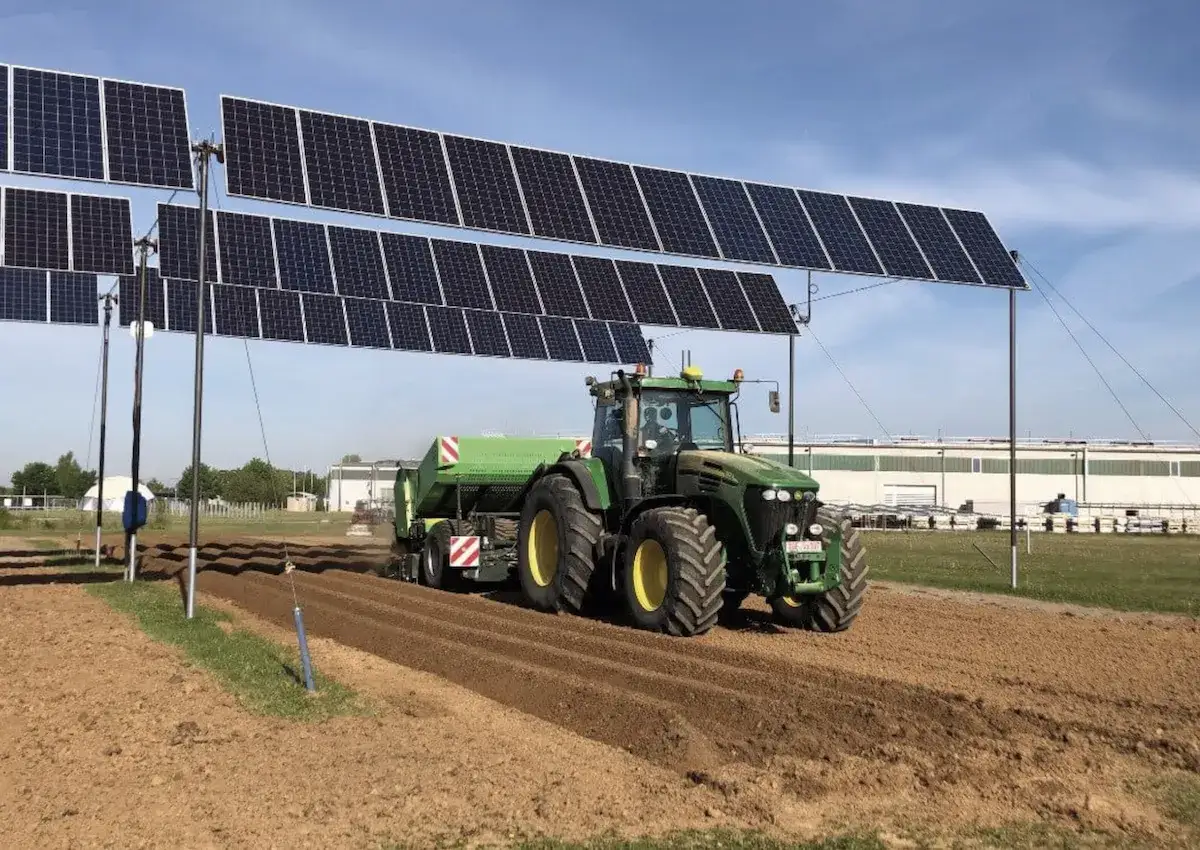 Trattore verde lavora un campo sotto un impianto agrivoltaico con pannelli solari sospesi.