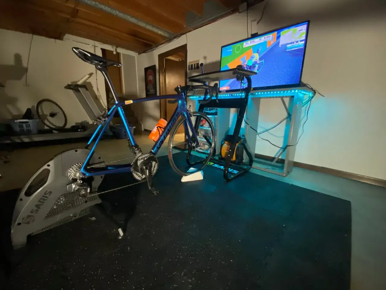 A blue road bike is set up on a Saris trainer, connected to a TV displaying a Zwift game. This indoor cycling setup includes various zwift compatible devices for an immersive workout.