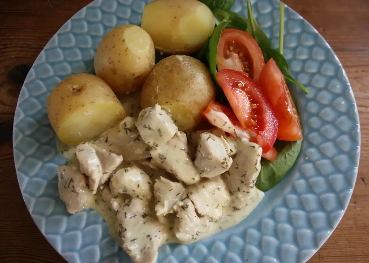 Kycklingbitar i kr&auml;mig citrons&aring;s med dill, serverade med kokt potatis, tomater och spenat p&aring; en bl&aring;m&ouml;nstrad tallrik.