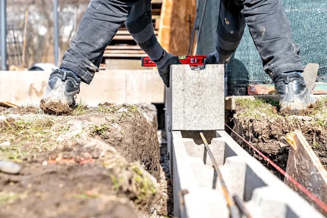 Budowa ściany fundamentowej z bloczków betonowych. Pracownik w rękawicach zakłada kolejny bloczek, sprawdzając poziom.