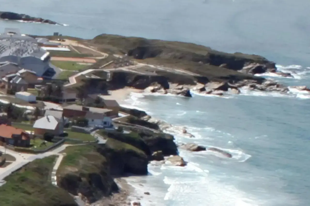 Vista aérea de casas y un complejo deportivo junto a la **playa de San Bartolo Barreiros**, con el mar agitado y rocas.