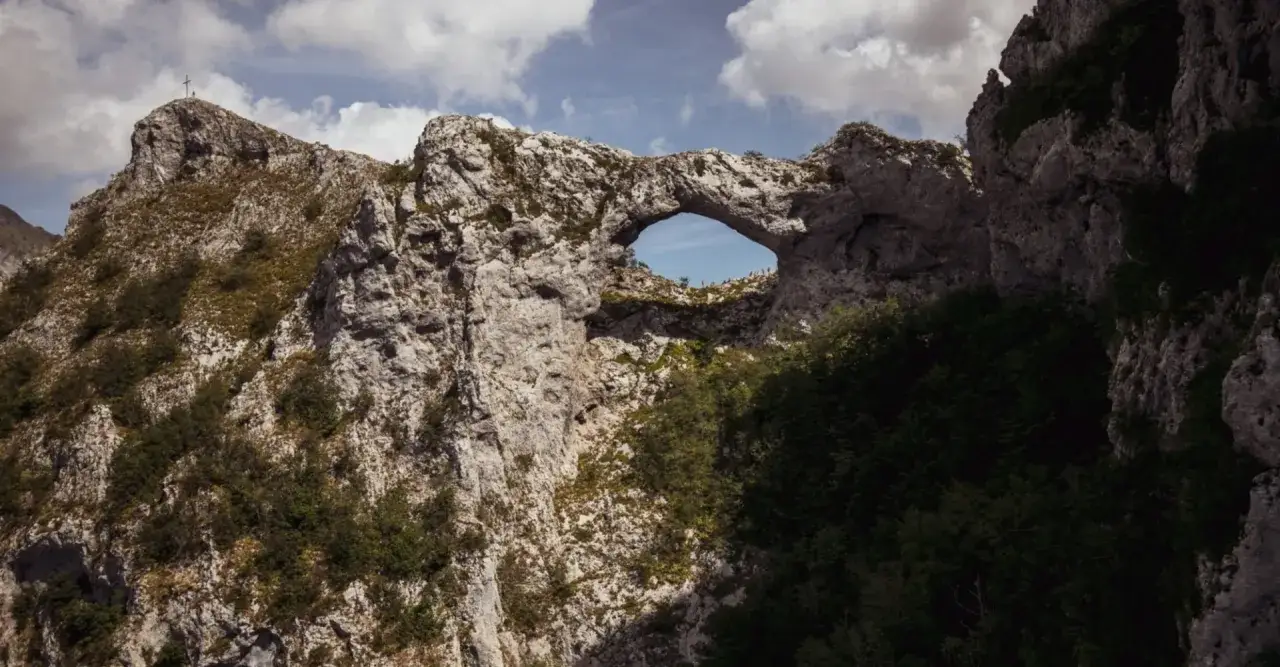 Il monte forato, un arco naturale tra rocce e vegetazione, domina il paesaggio. In cima, una croce segna la vetta.