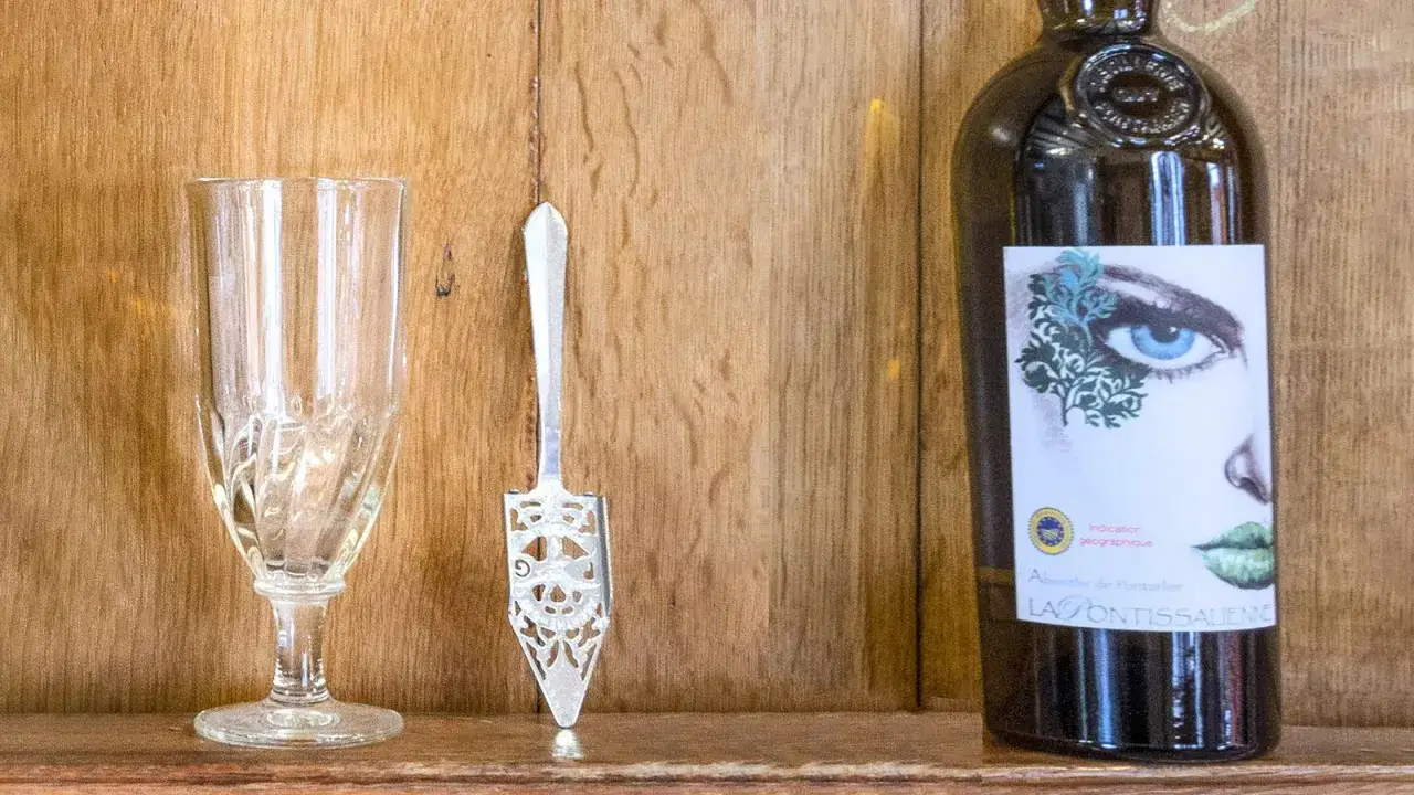 Ein Glas, ein Löffel und eine Flasche Absinth. Die Flasche zeigt ein Auge und Lippen. Die Absinth Wirkung wird oft als berauschend beschrieben.
